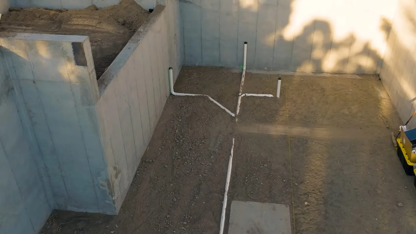 Bare basement floor with white PVC drain lines laid across the ground inside block walls